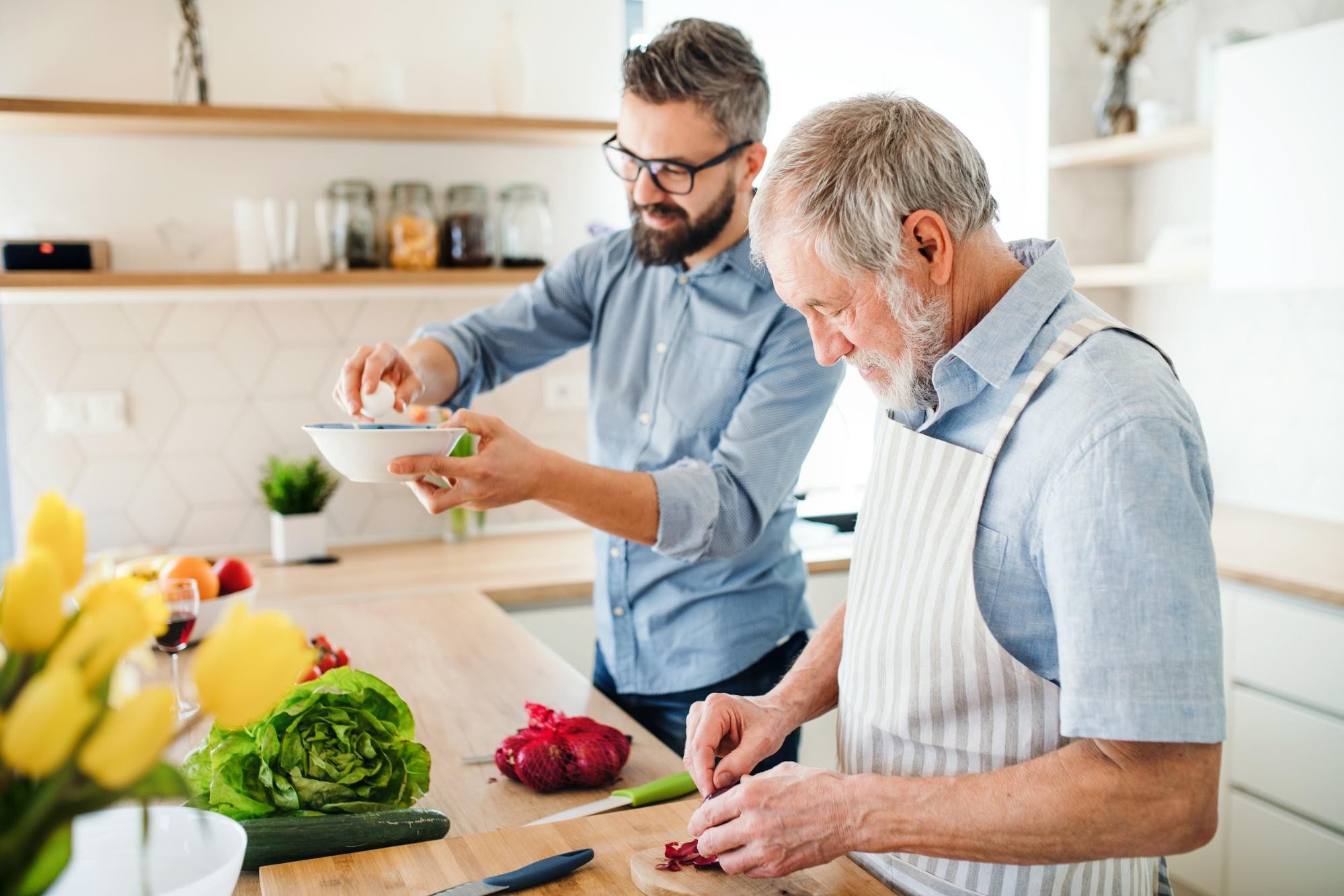 Older man chopping vegetables and younger man holding bowl cooking together in modern kitchen