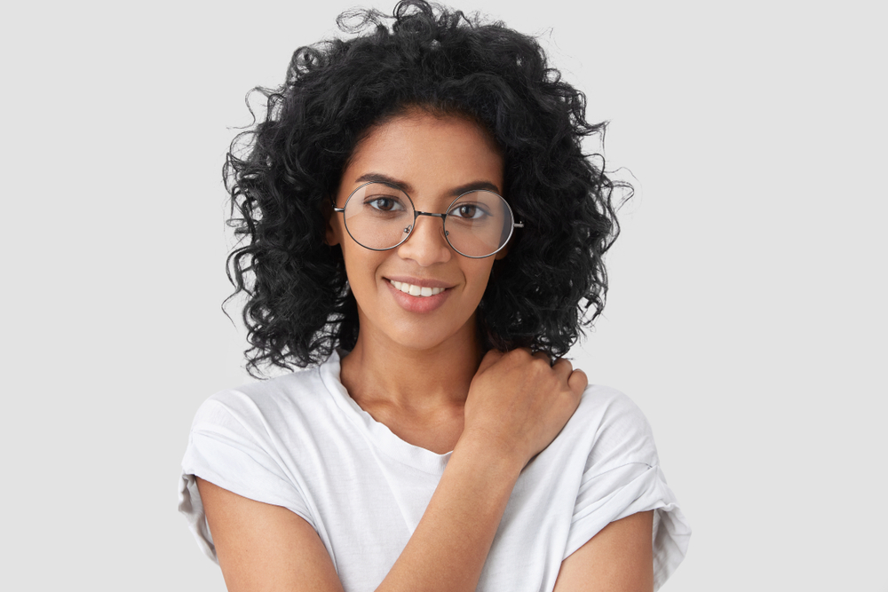 Woman with curly black hair and glasses smiling in white t-shirt against gray background