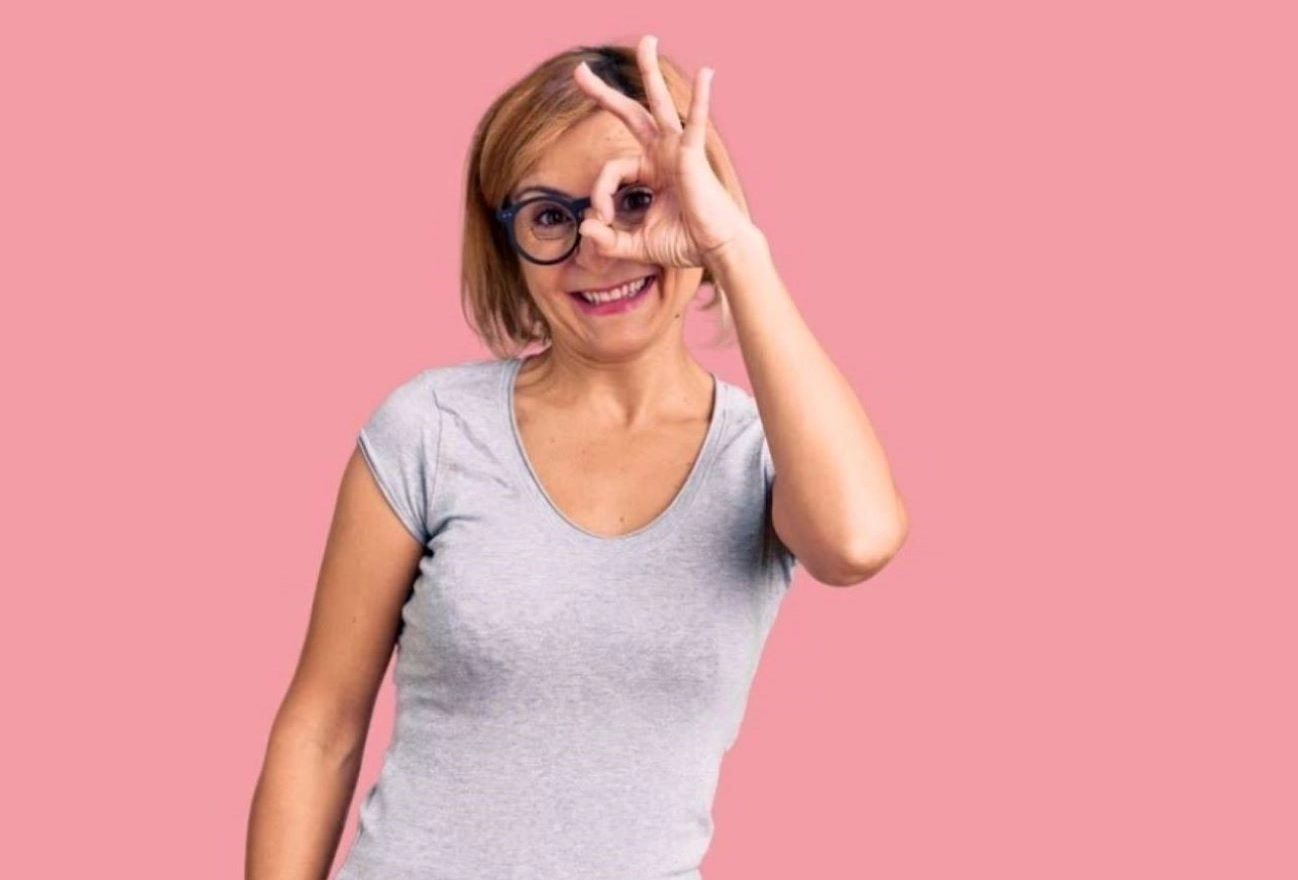 Woman in glasses and gray t-shirt making OK gesture with hand against pink background