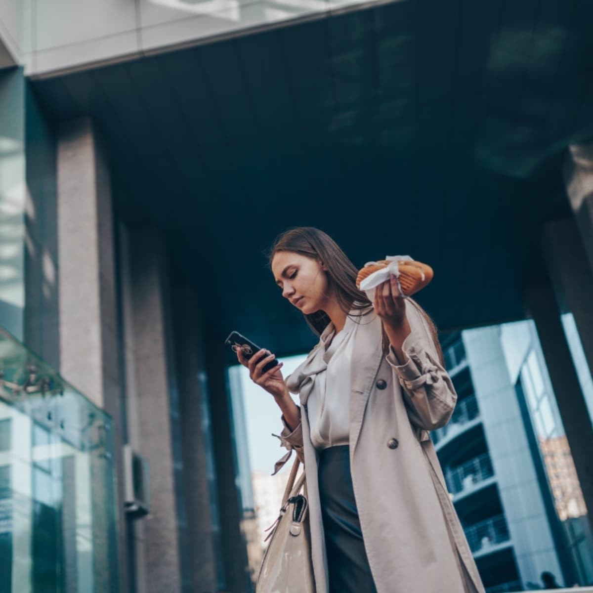 Woman in trench coat holding croissant and checking phone on city street