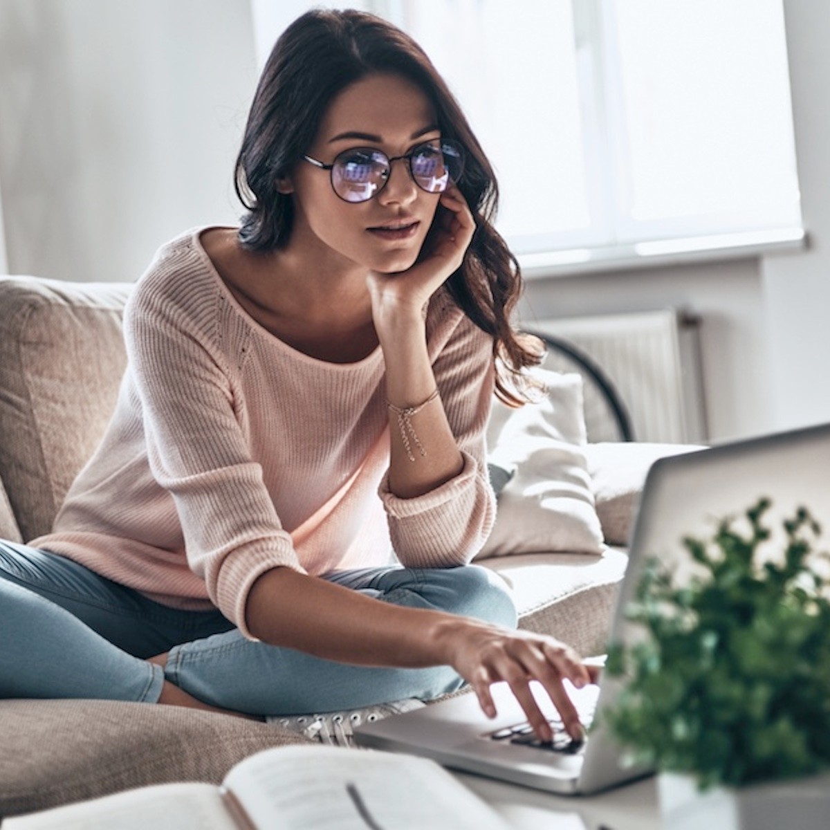 Woman in pink sweater wearing glasses working on laptop while sitting on couch in bright modern workspace