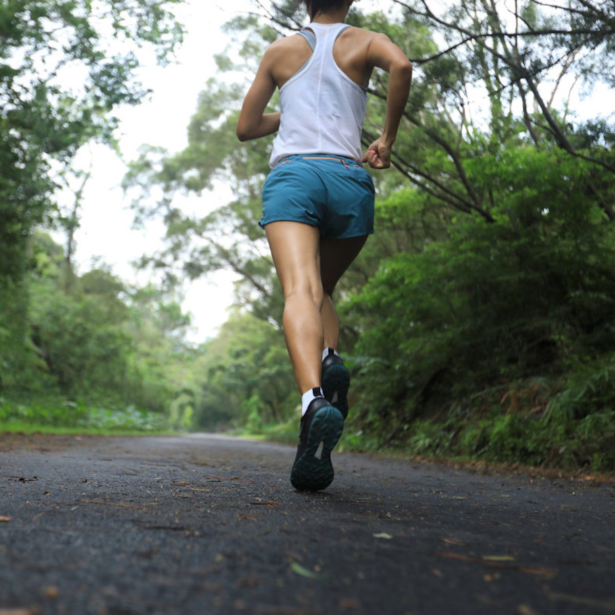 Person jogging on tree-lined asphalt road wearing white tank top and blue shorts