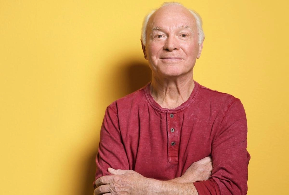 Older man with white hair in burgundy henley shirt, arms crossed, against yellow background