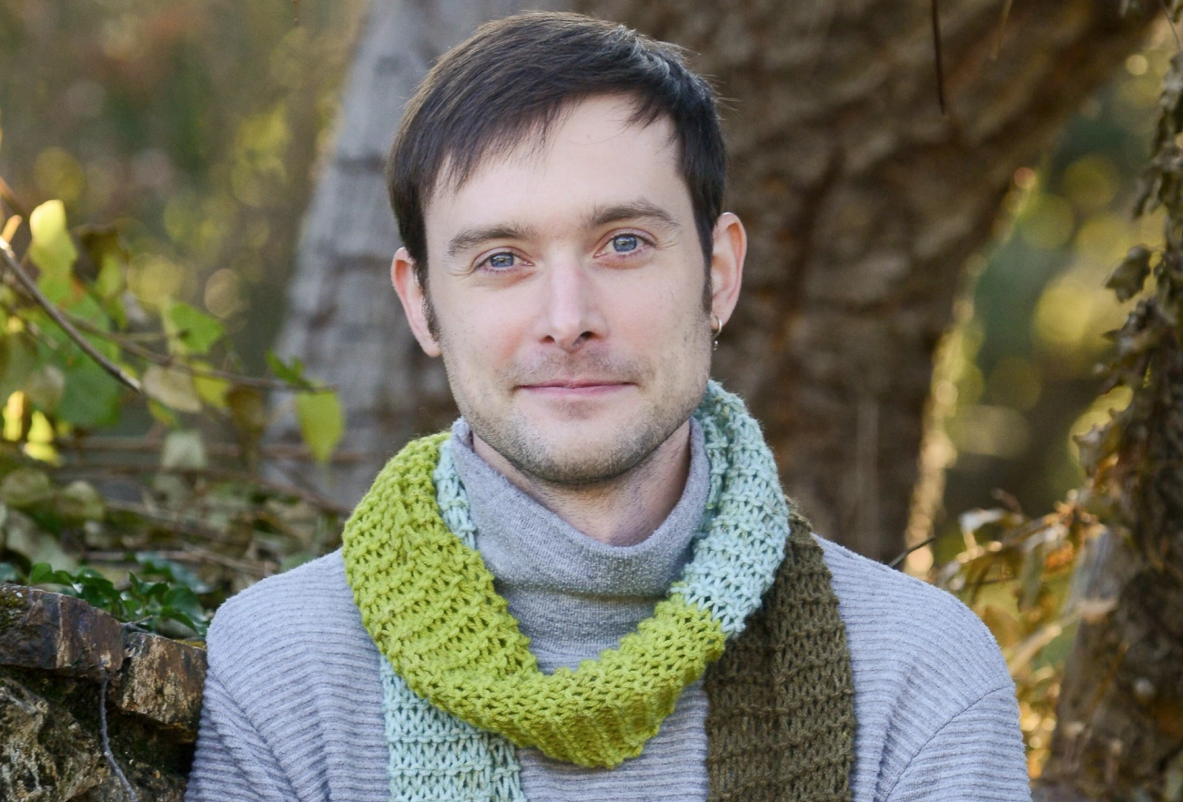 Man in gray sweater and green knit scarf against ivy-covered stone wall outdoors