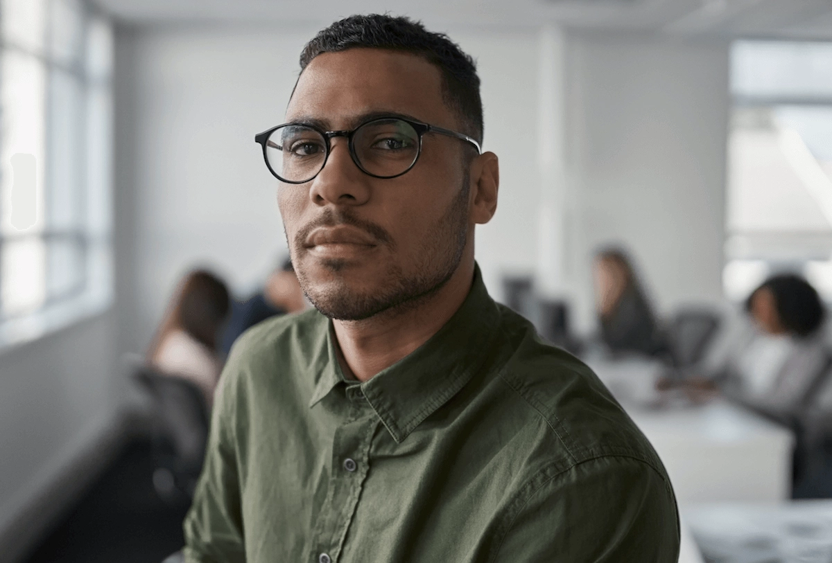 Man wearing glasses and olive shirt in office setting with blurred colleagues in background