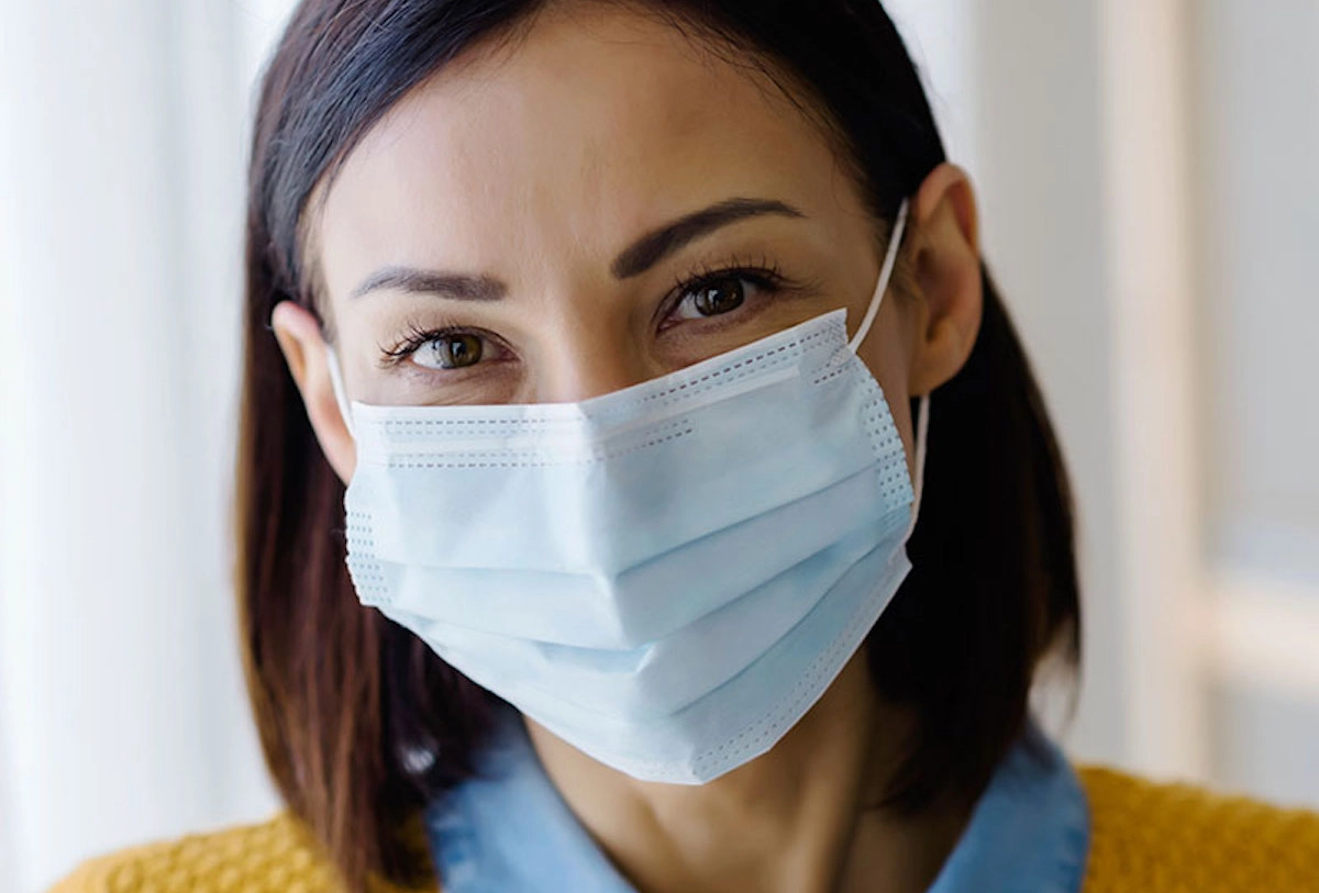 Person wearing a white surgical mask and yellow top against a blurred indoor background