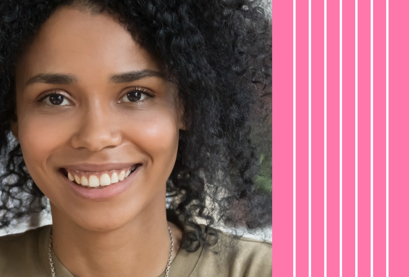 Smiling person with curly dark hair wearing a tan shirt and necklace against a pink striped background