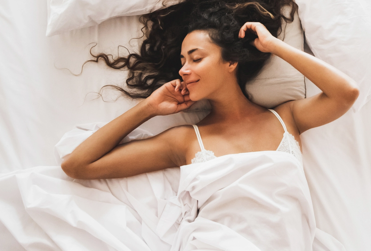 Woman sleeping peacefully in white bed with hand resting on face and spread dark hair on pillow