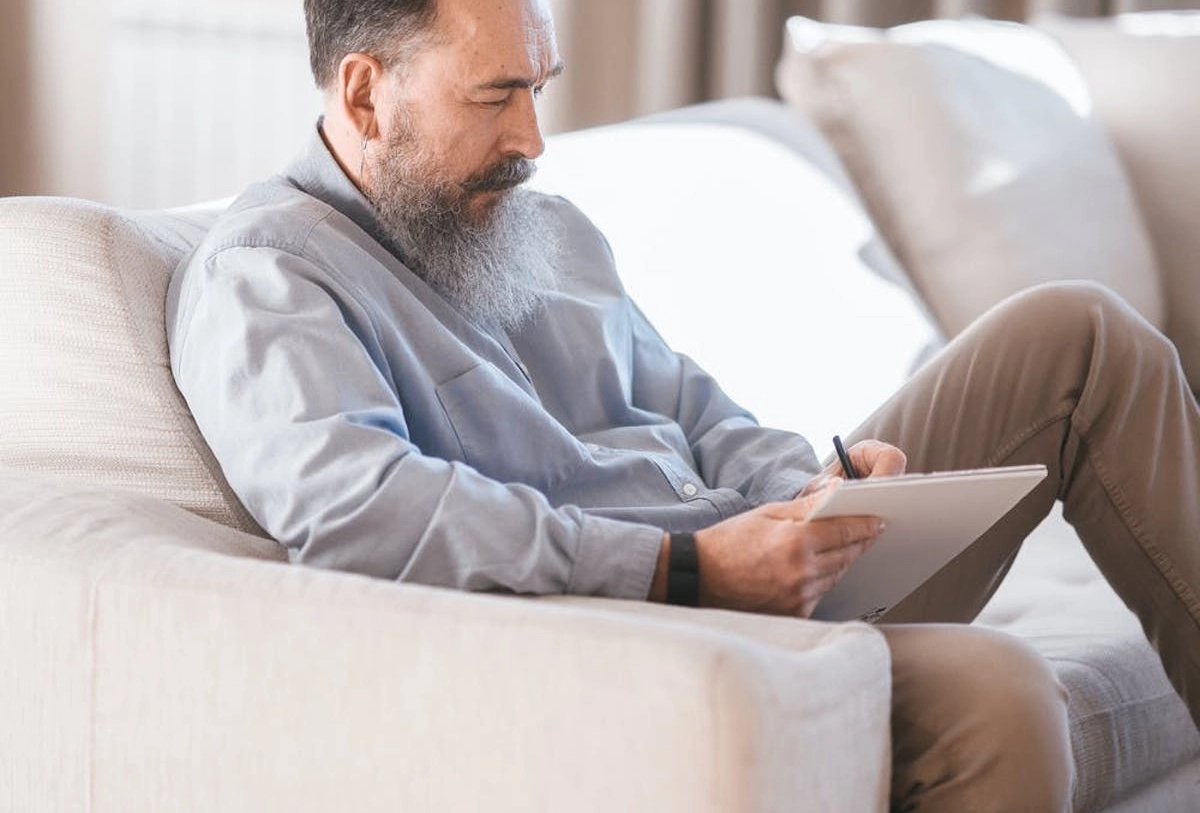 Older man sitting on couch, holding tablet and stylus, looking down thoughtfully while surrounded by white pillows