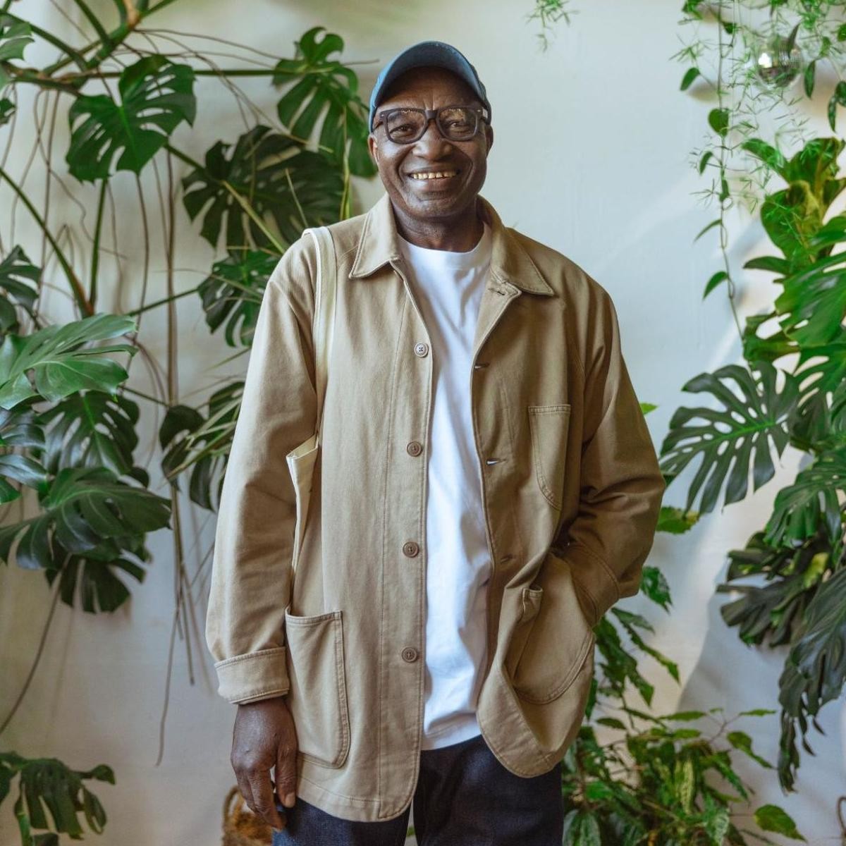 Smiling man in beige jacket and blue cap standing among potted green plants indoors