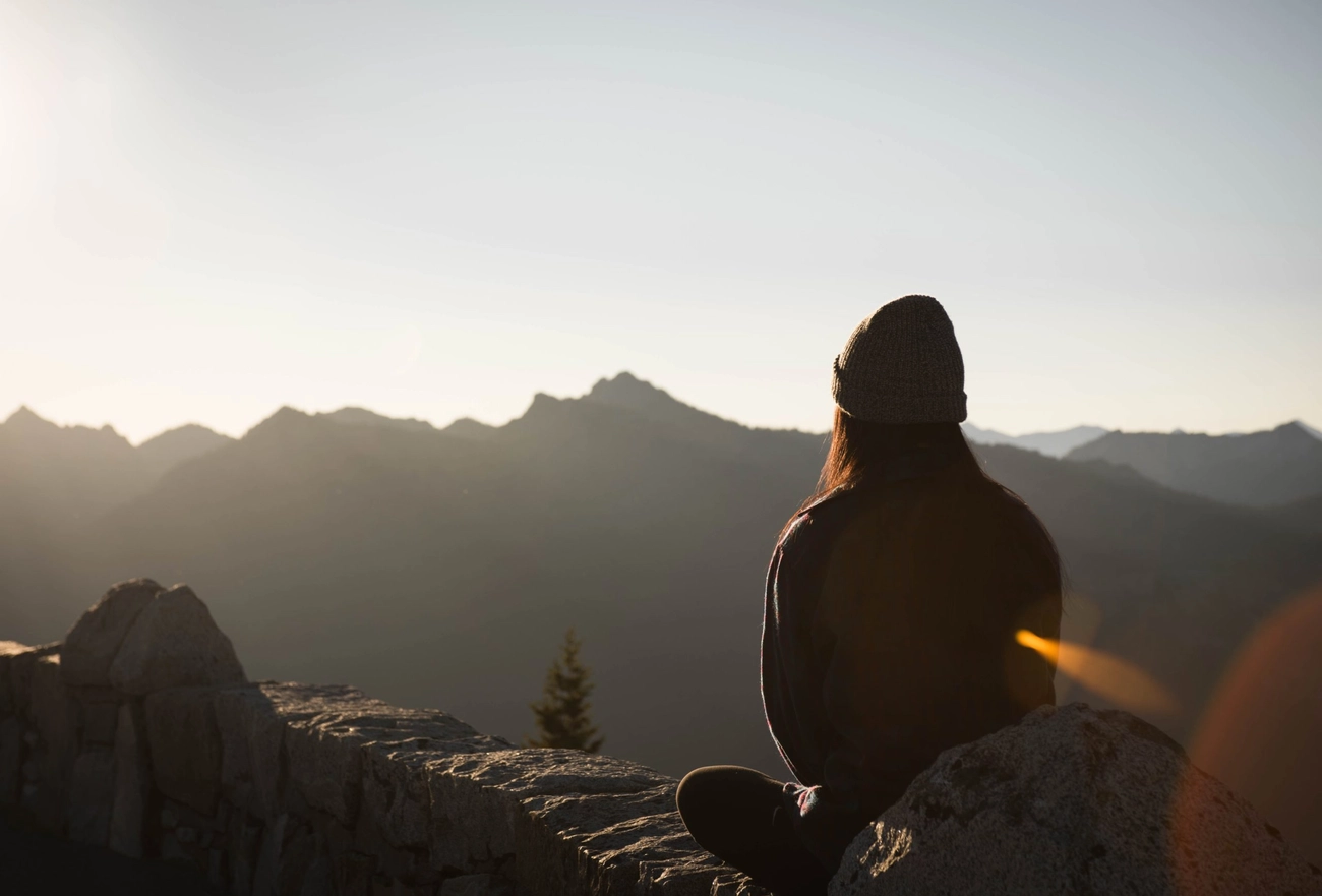Woman sitting silhouetted on a mountain peak overlooking vast mountain range at sunset