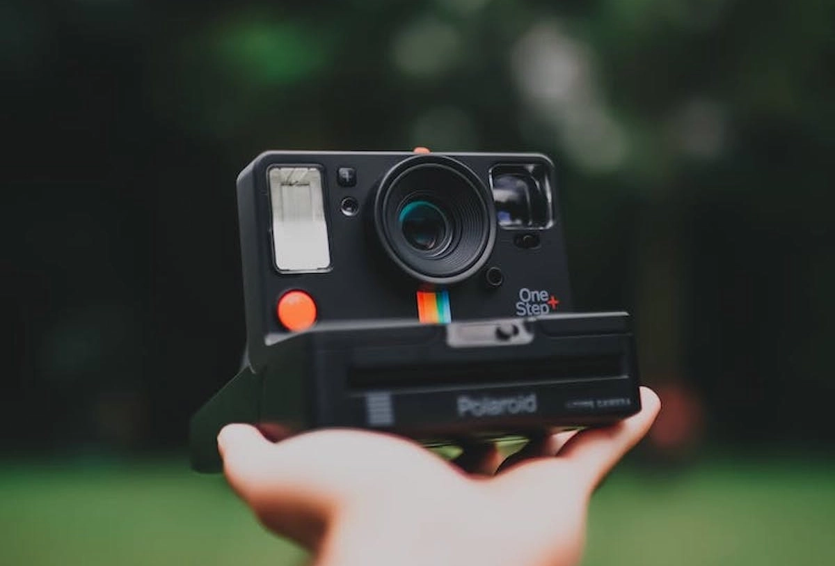 Hand holding black Polaroid OneStep instant camera with orange button and rainbow stripe against blurred green background