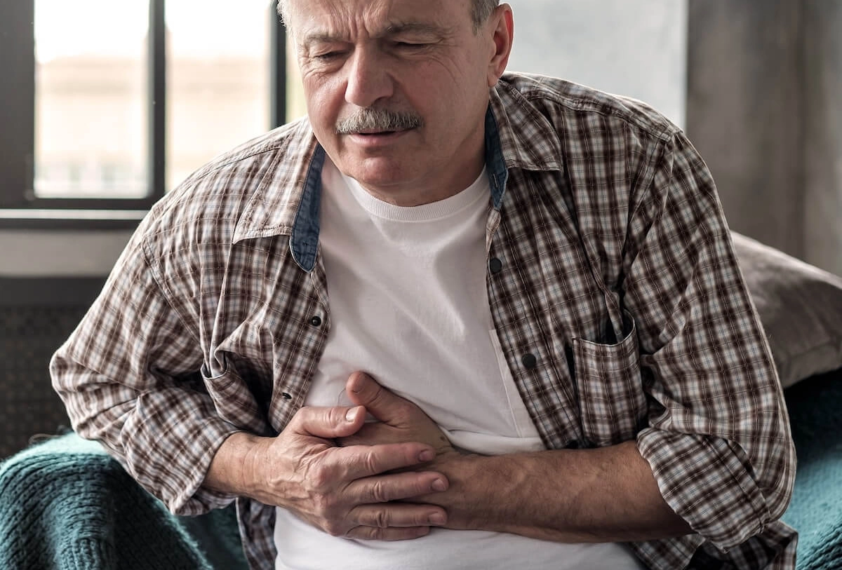 Older man in plaid shirt clutching stomach in pain while sitting on couch by window