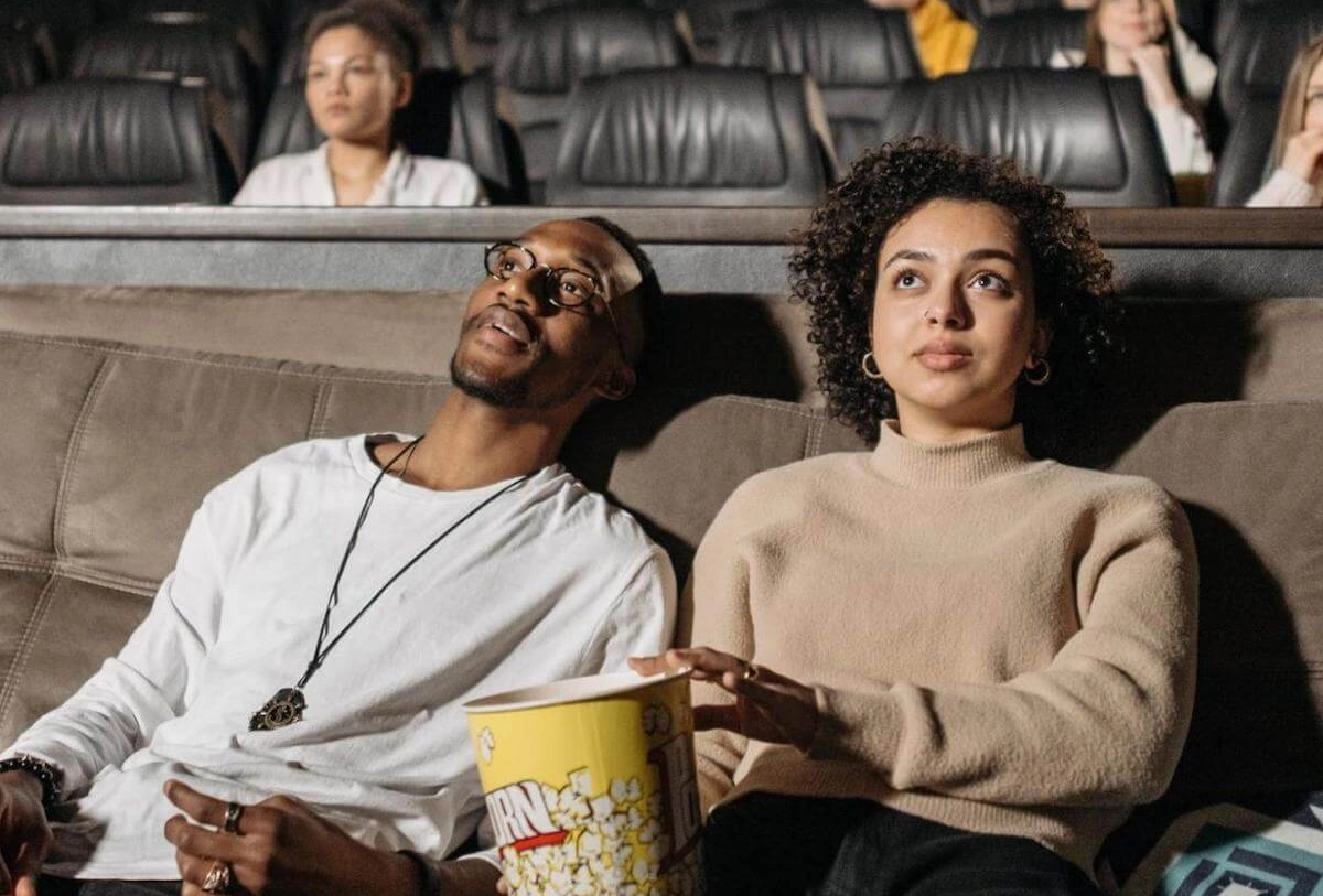 Couple watching movie in theater holding popcorn, other patrons seated in background