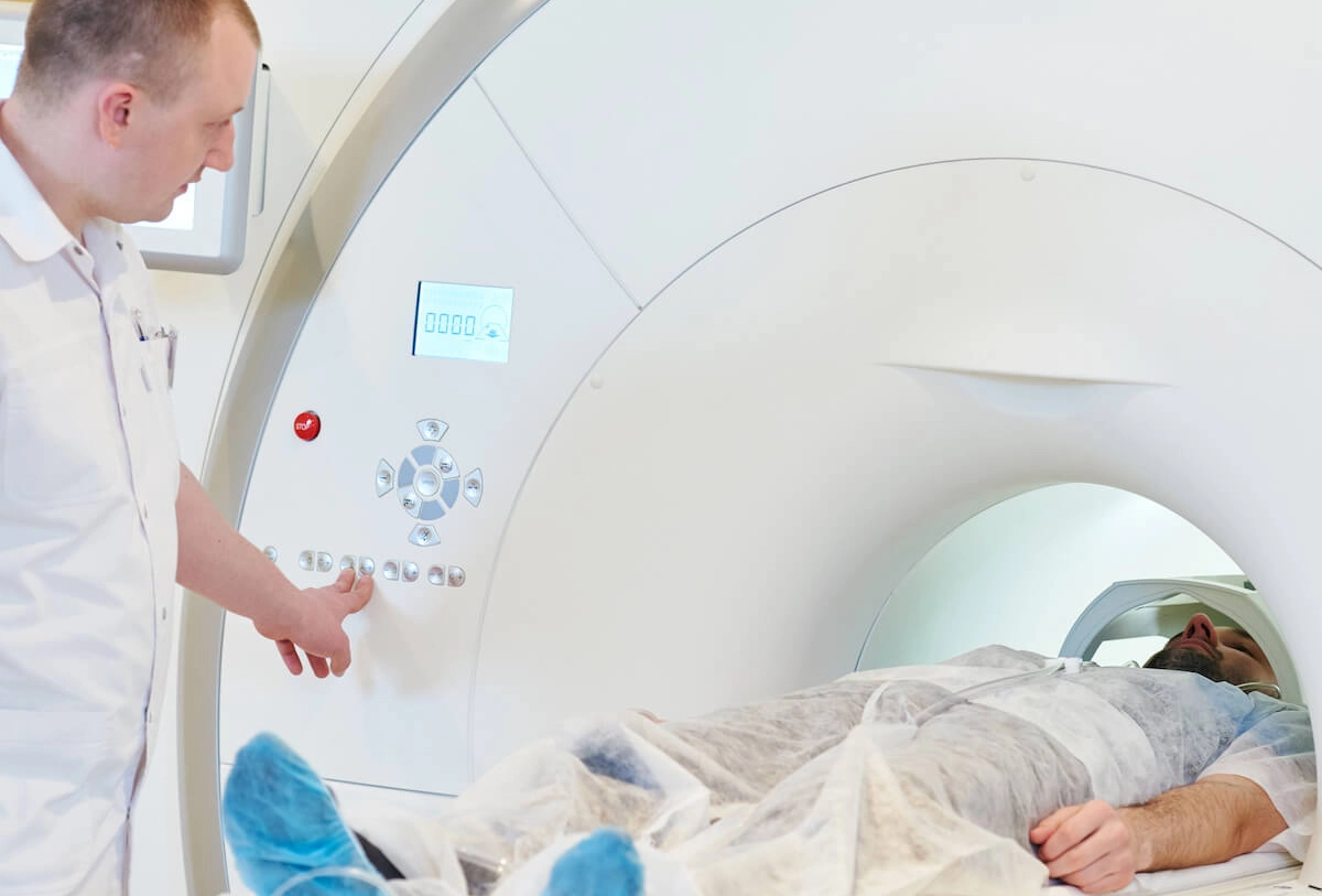Healthcare worker standing at CT scanner control panel while patient undergoes medical imaging scan inside machine