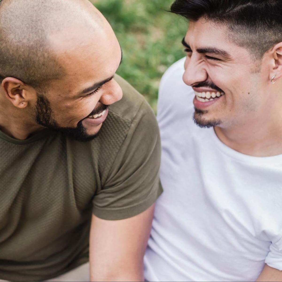 Two men smiling and laughing together outdoors, one in olive shirt and one in lavender shirt, with green foliage behind