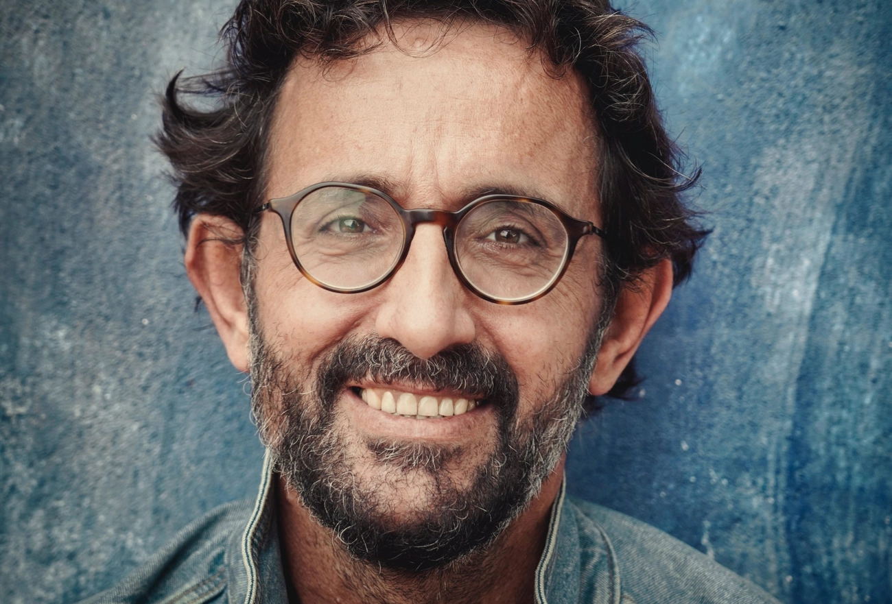 Middle-aged man with beard and round glasses smiling at camera against blue background