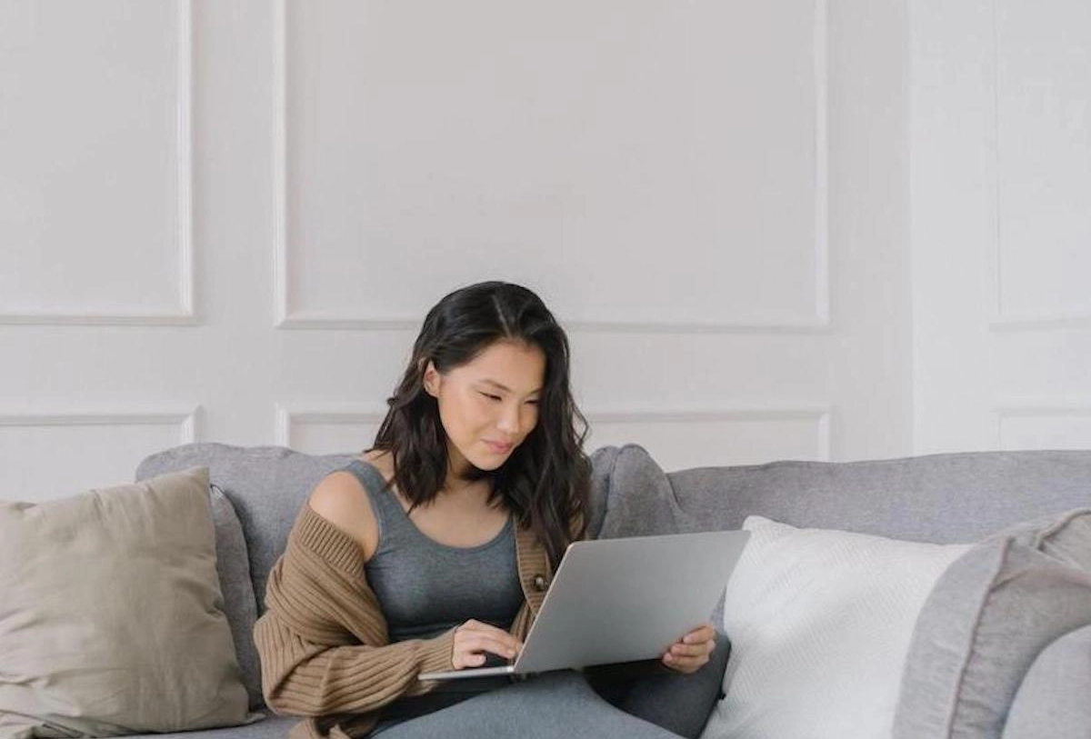 Woman using laptop on gray couch with pillows in bright living room