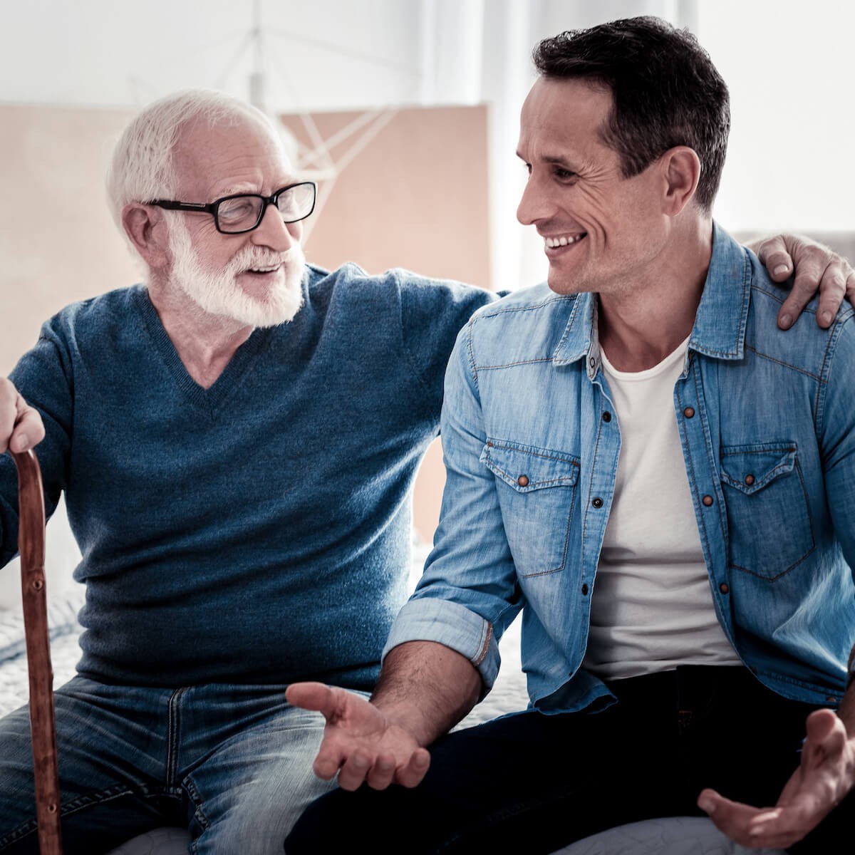 Elderly man in glasses and sweater with arm around younger man in denim, both smiling warmly together