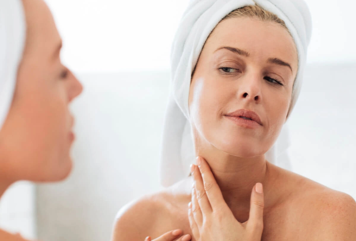 Woman in white towel examining her face in mirror, touching jawline while checking skin