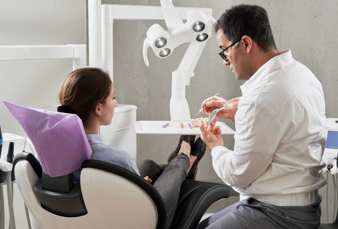 Dentist in white coat explaining dental treatment to female patient in purple bib in modern dental office with overhead light