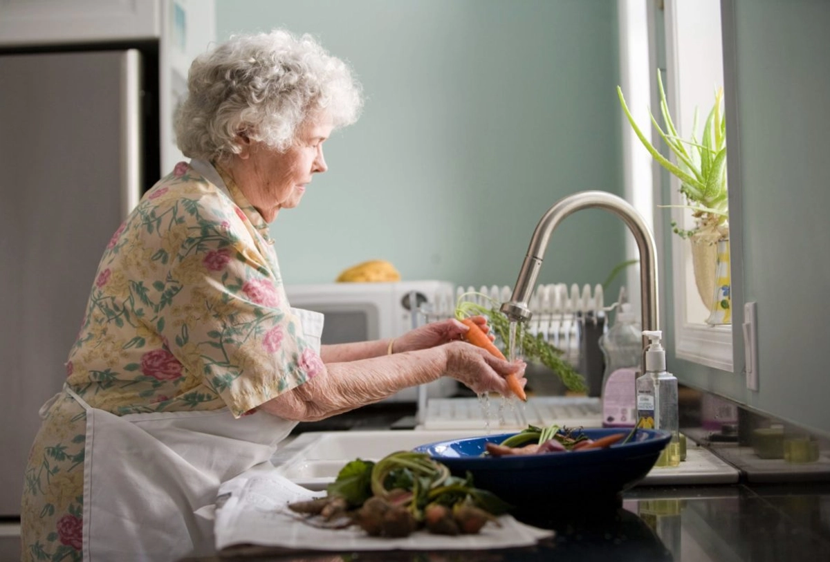 Elderly woman with white hair washing vegetables at kitchen sink in a modern kitchen with fresh produce on counter