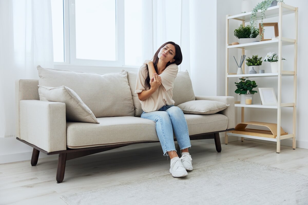 Woman holding neck in pain while sitting on sofa in bright, minimalist living room with plants