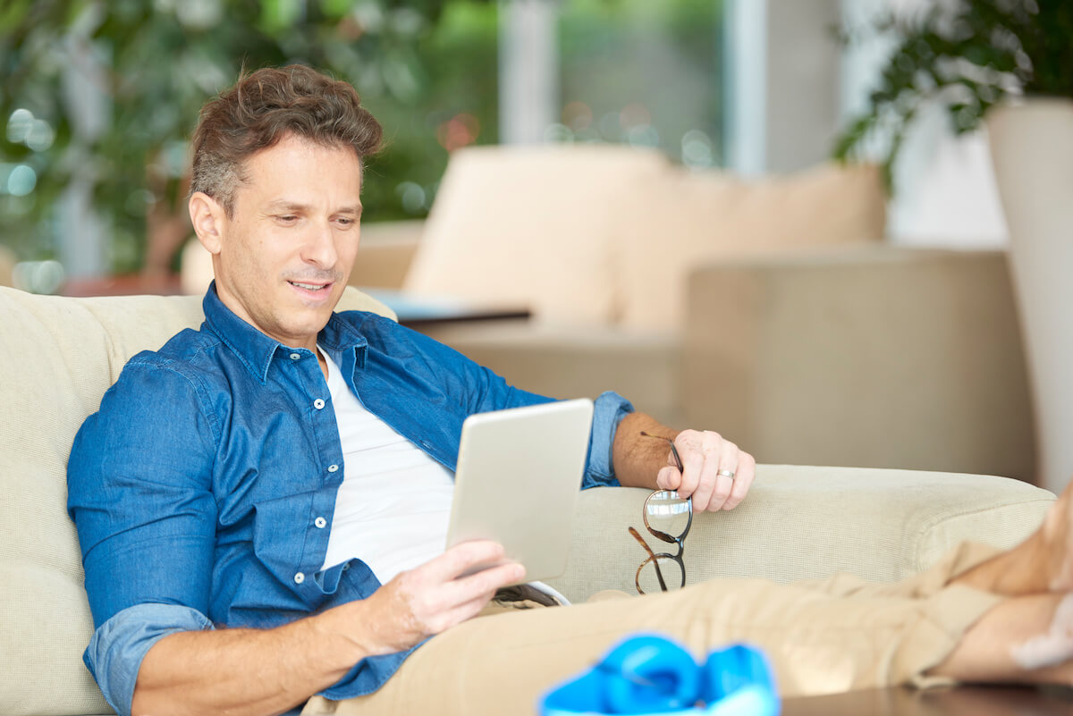 Man sitting on couch using tablet with glasses and blue headphones in modern living room with plants