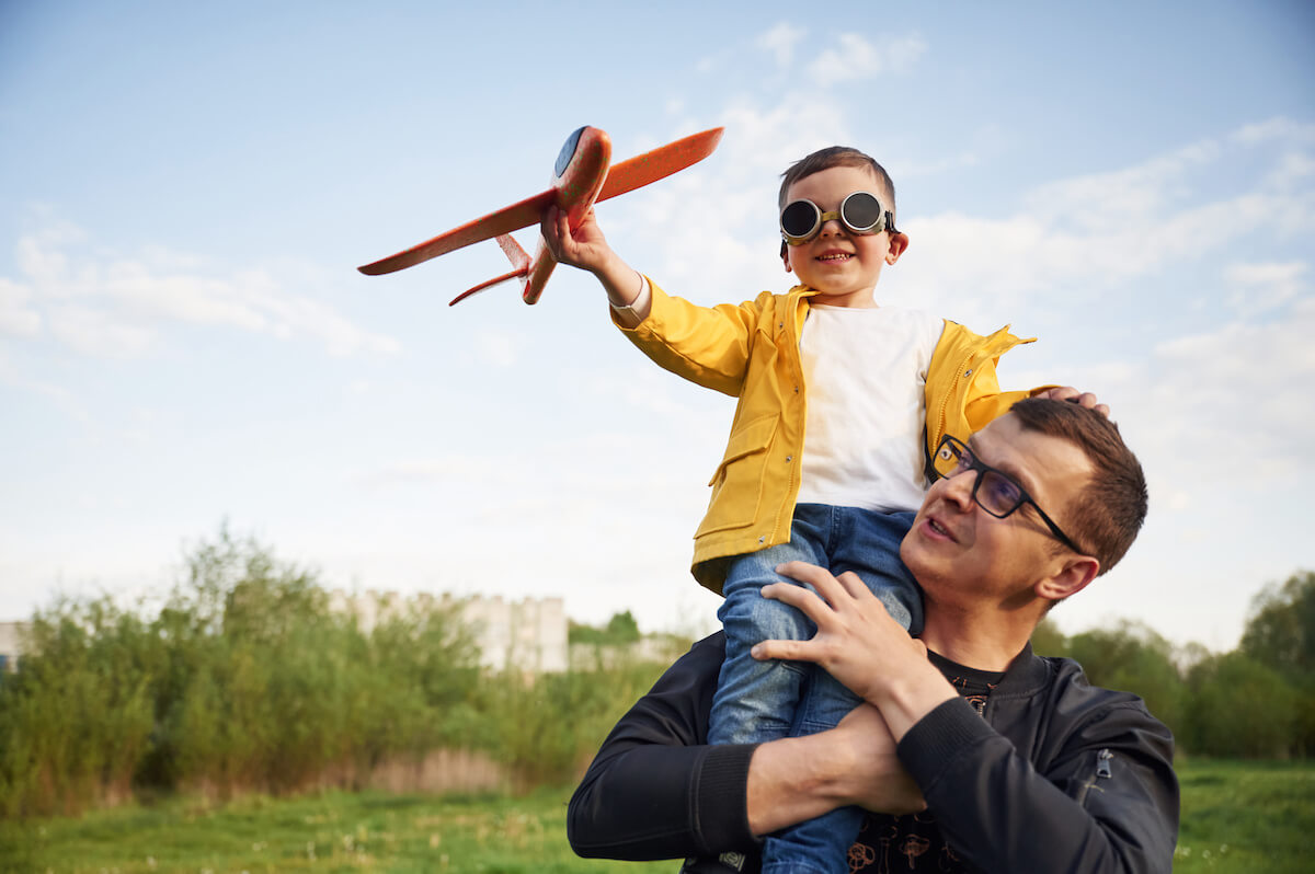 Father holding son on shoulders playing with red toy airplane in park with green landscape