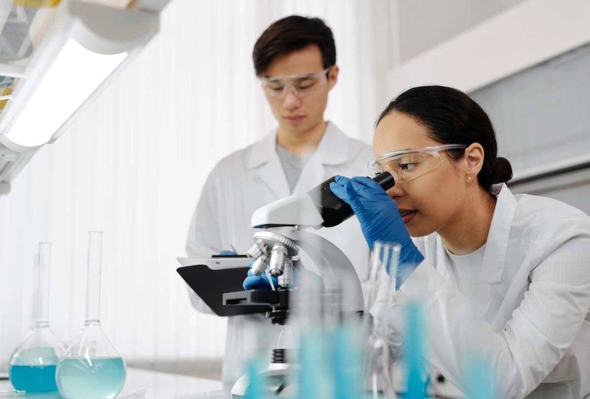 Female scientist examining specimen under microscope in laboratory with colleague in background