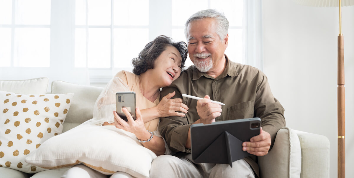 Smiling mature couple on couch looking at smartphone and tablet together in bright living room