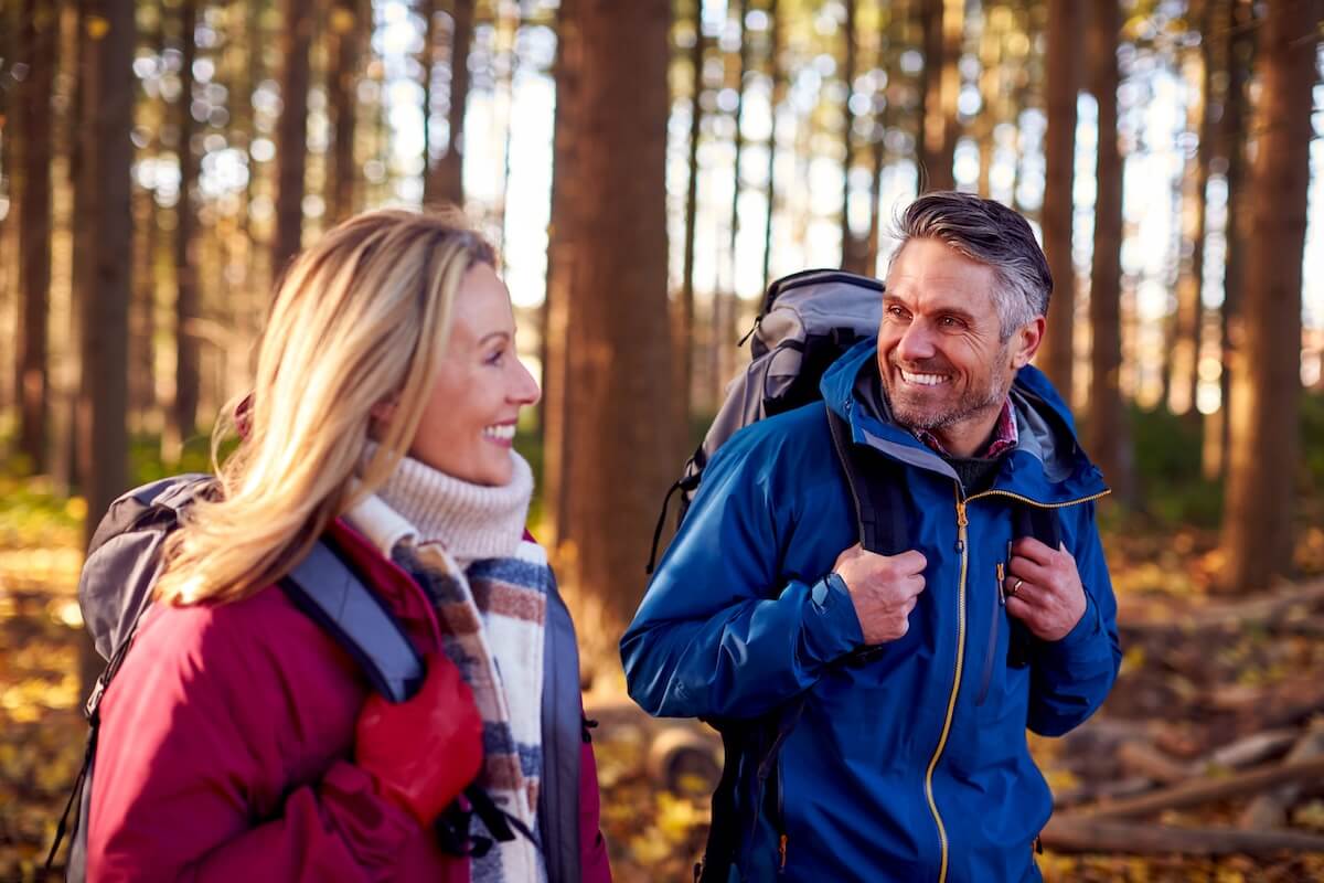 Couple hiking together on a forest trail wearing backpacks and smiling in autumn