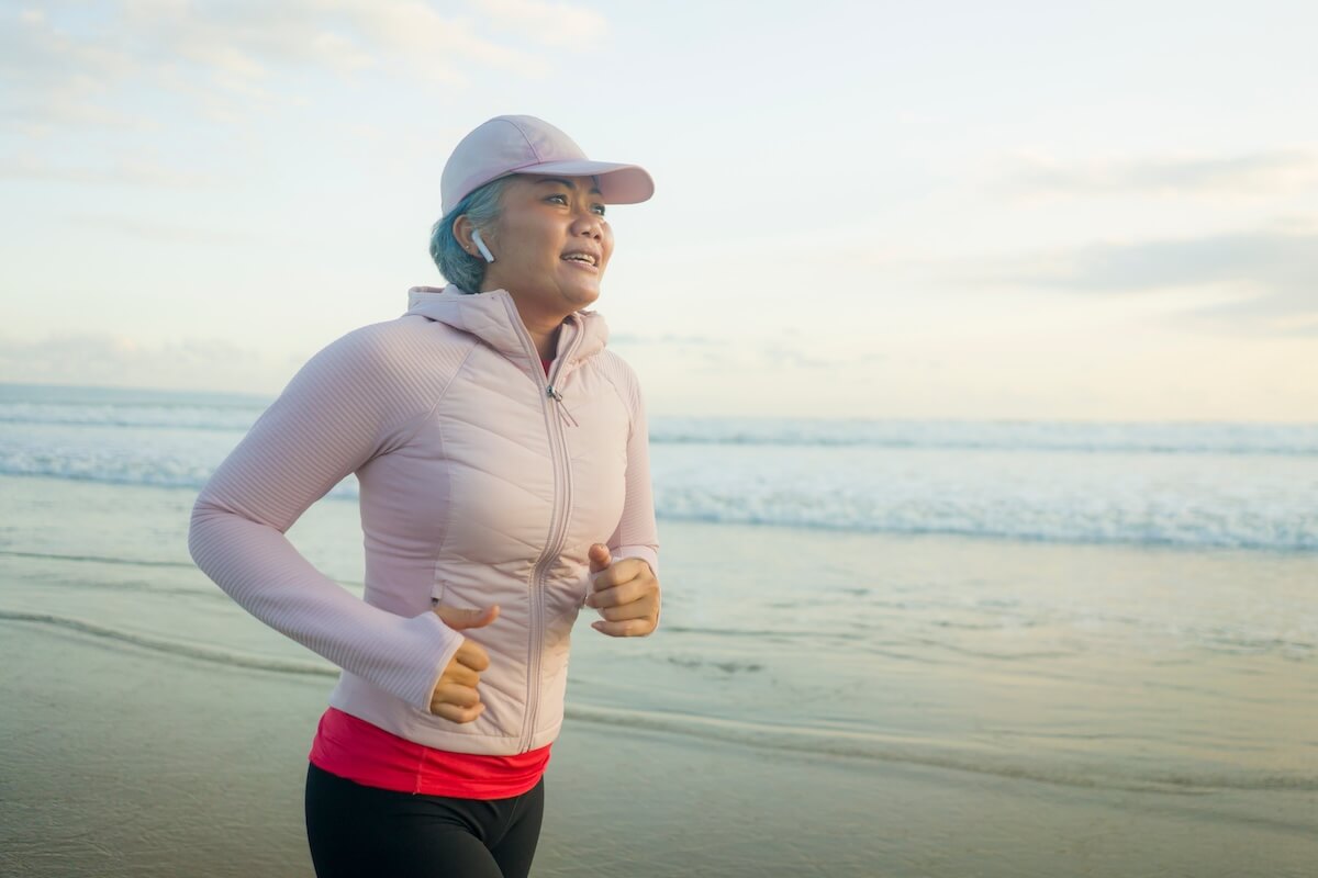 Woman jogging on beach wearing pink jacket and cap, ocean in background