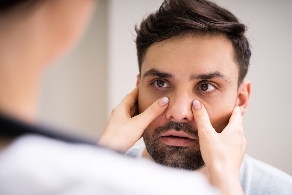 Person examining sinus area or under-eye region in mirror, touching face with fingers while checking for discomfort