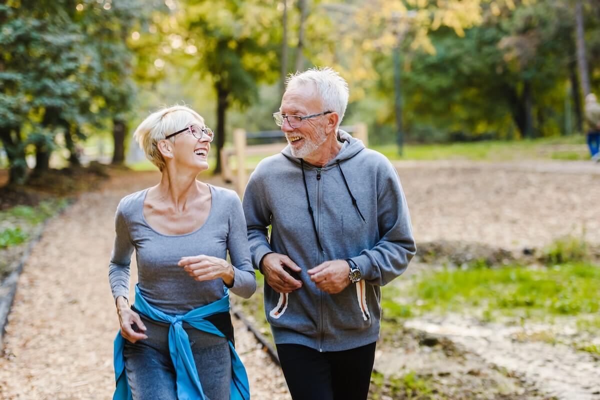 Senior couple laughing while walking together on nature trail, both in gray athletic wear with autumn trees in background