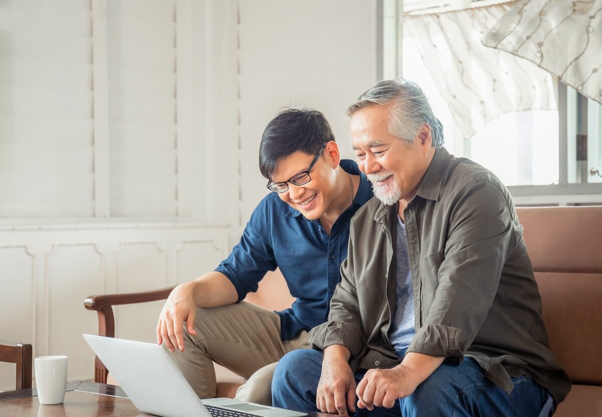 Younger and older man sharing a laptop moment on couch, both smiling while viewing screen in bright home interior