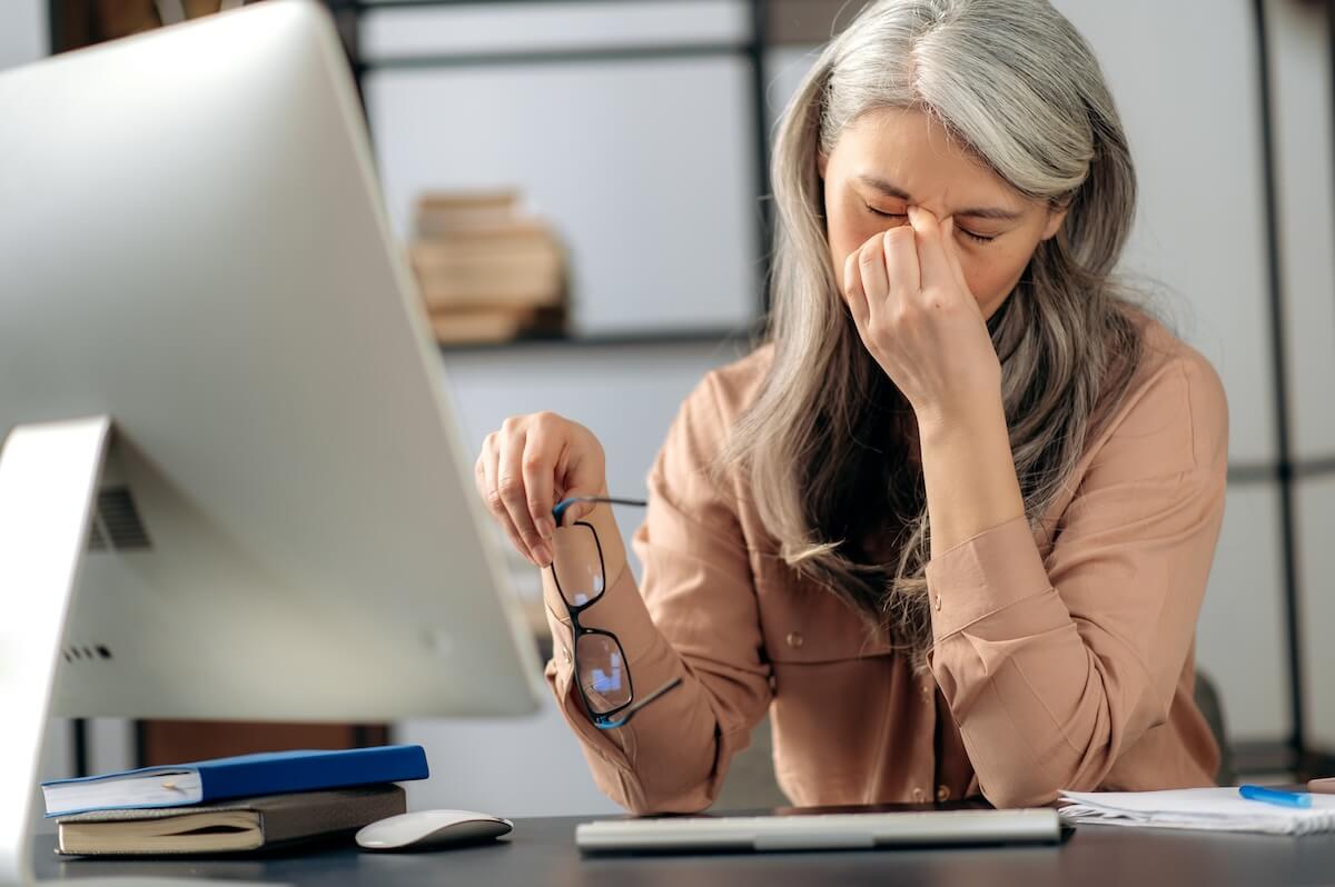 Person with silver hair experiencing eye strain at computer desk, rubbing nose bridge while holding glasses in office setting