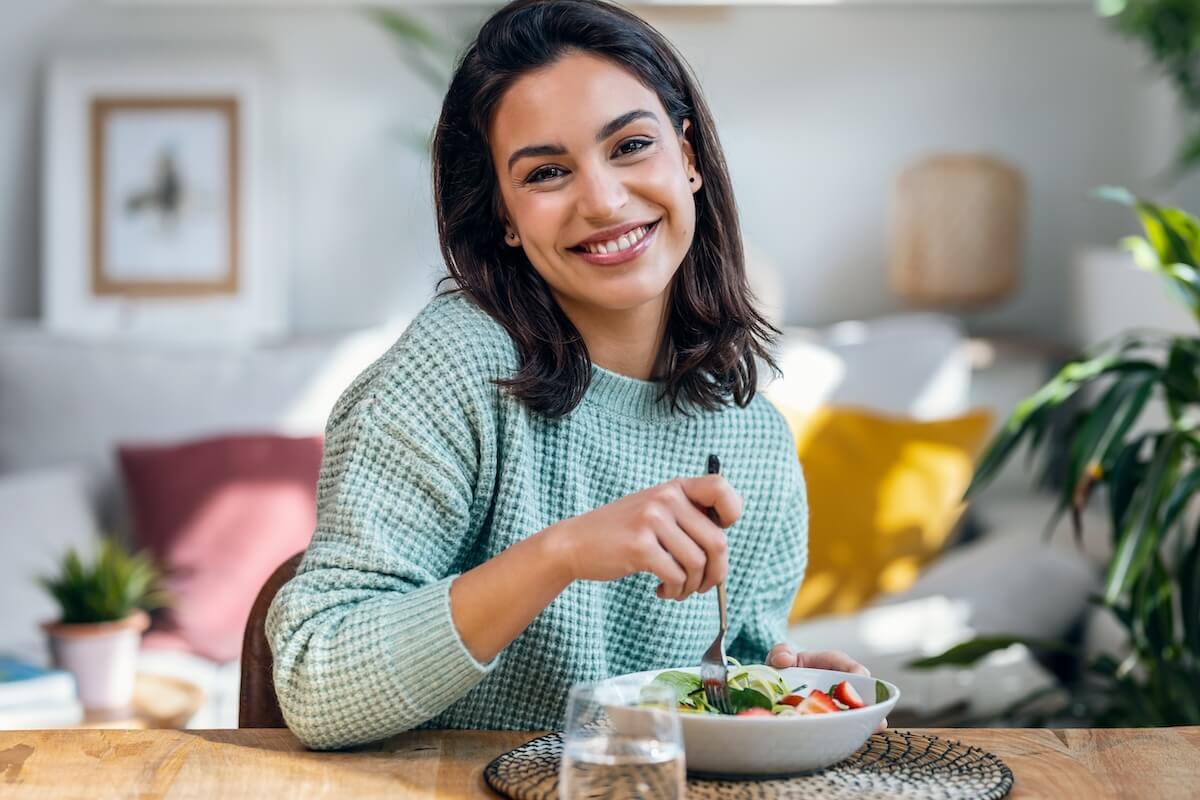 Person in mint sweater enjoying a fresh salad at wooden table in bright home interior with plants and colorful decor