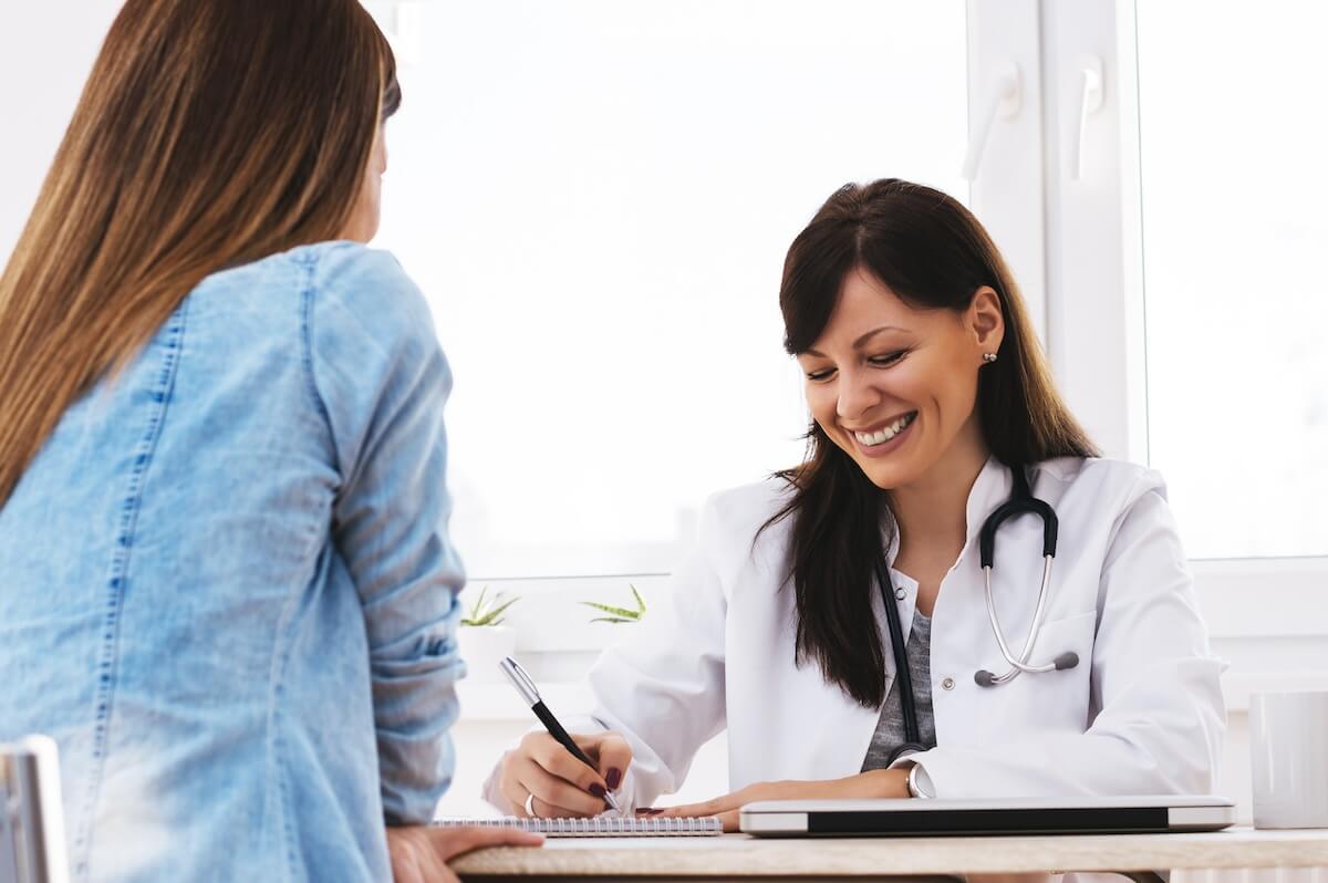 Doctor with stethoscope taking notes during patient consultation in bright medical office with patient in denim shirt