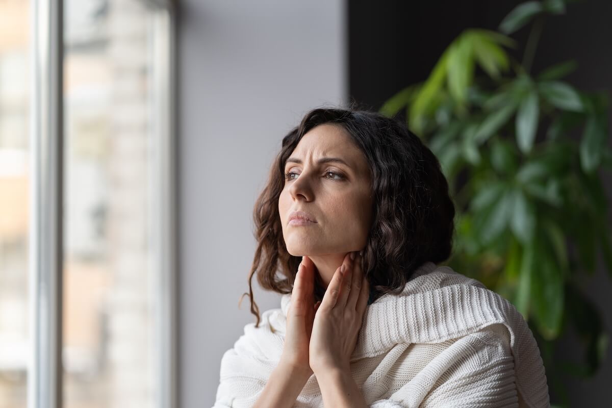 Person touching throat with concerned expression by window, white sweater, houseplant in background