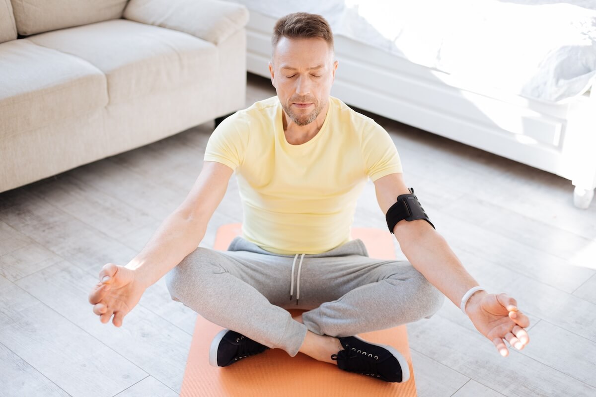Person in yellow shirt meditating cross-legged on orange yoga mat at home, wearing fitness tracker and gray sweatpants