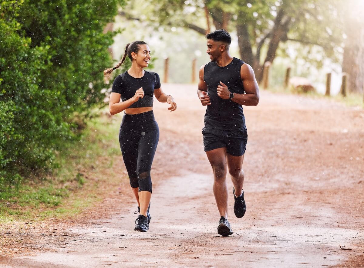 Two people jogging together on a forest path, wearing black athletic wear and smiling at each other in golden sunlight