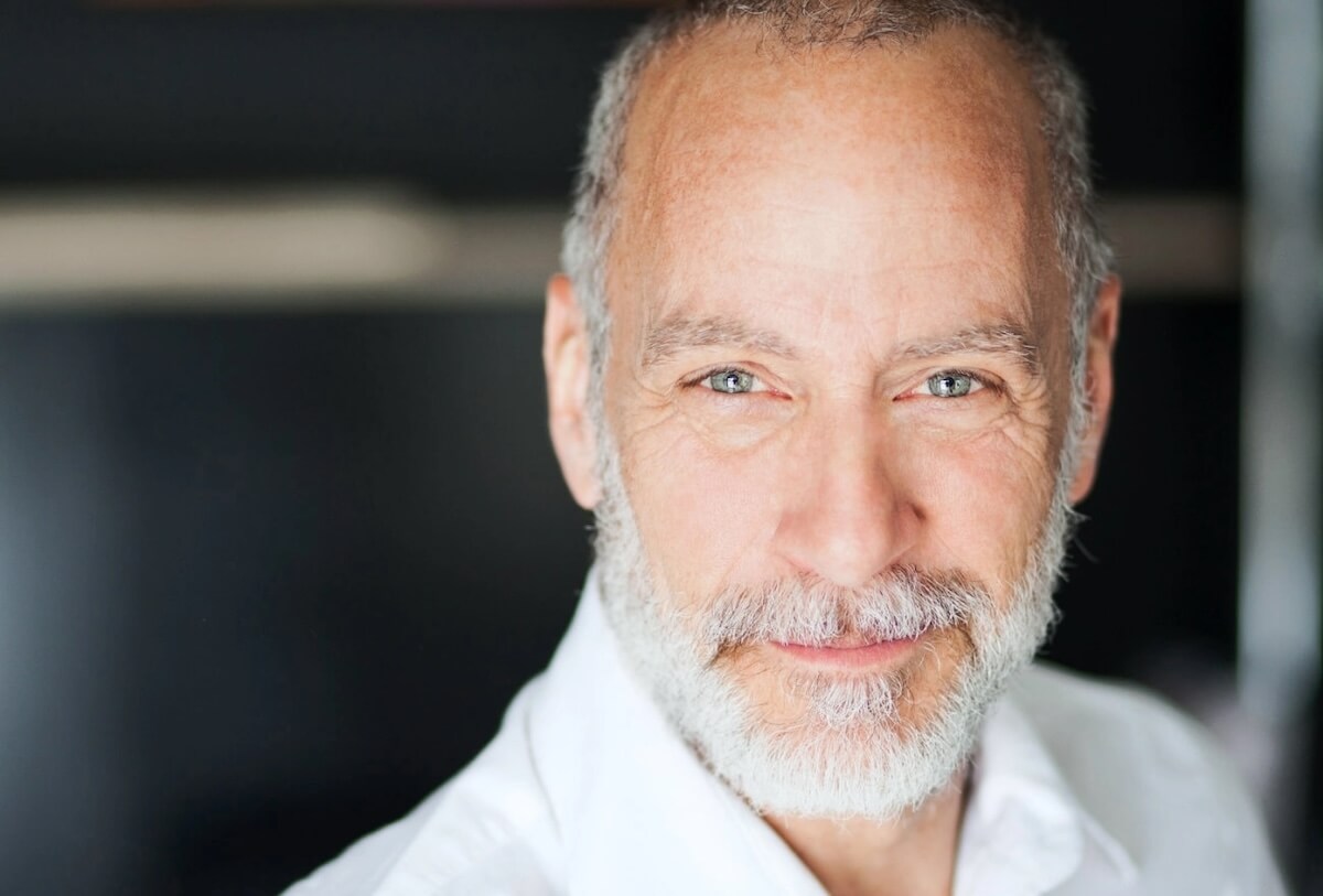 Older man with gray beard wearing white shirt against blurred background