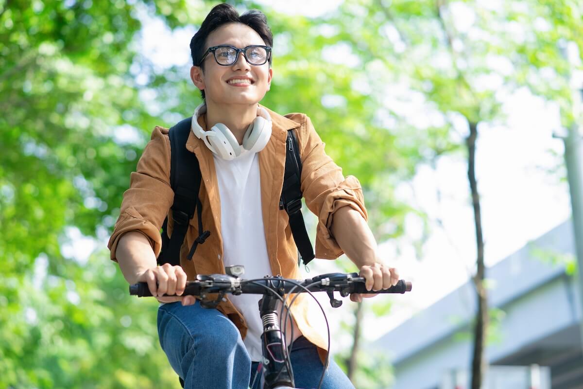 Young man in glasses and brown jacket riding bicycle outdoors with headphones around neck and green foliage background