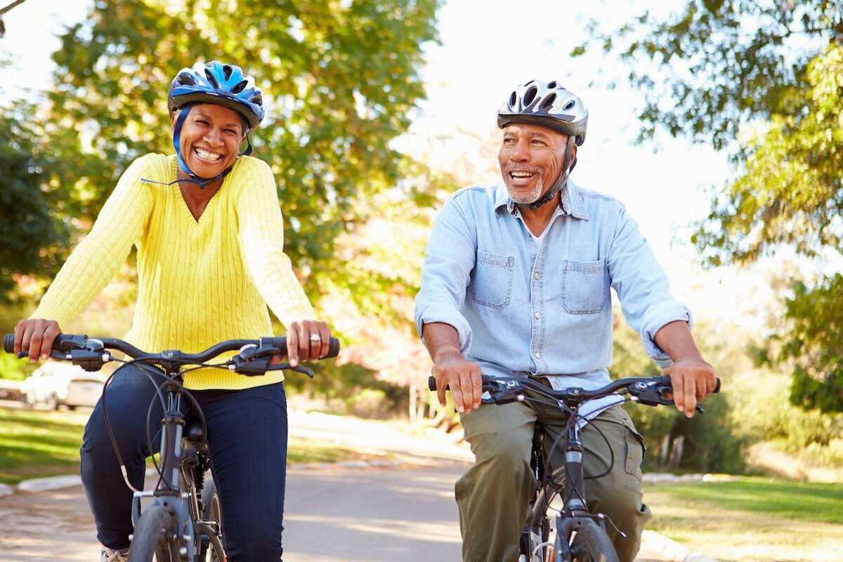 Senior couple cycling together on park path, wearing helmets, yellow and blue tops, smiling on sunny day