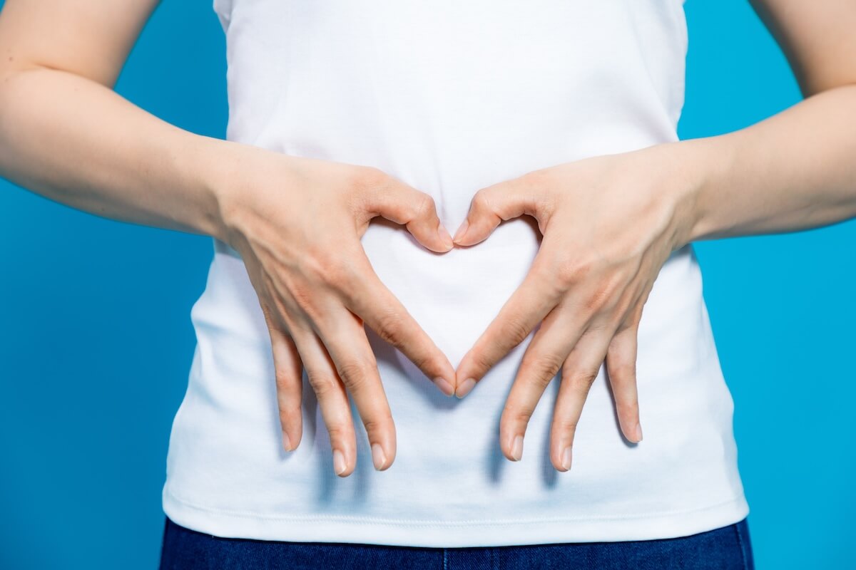 Person making heart shape with hands over abdomen, wearing white shirt against blue background