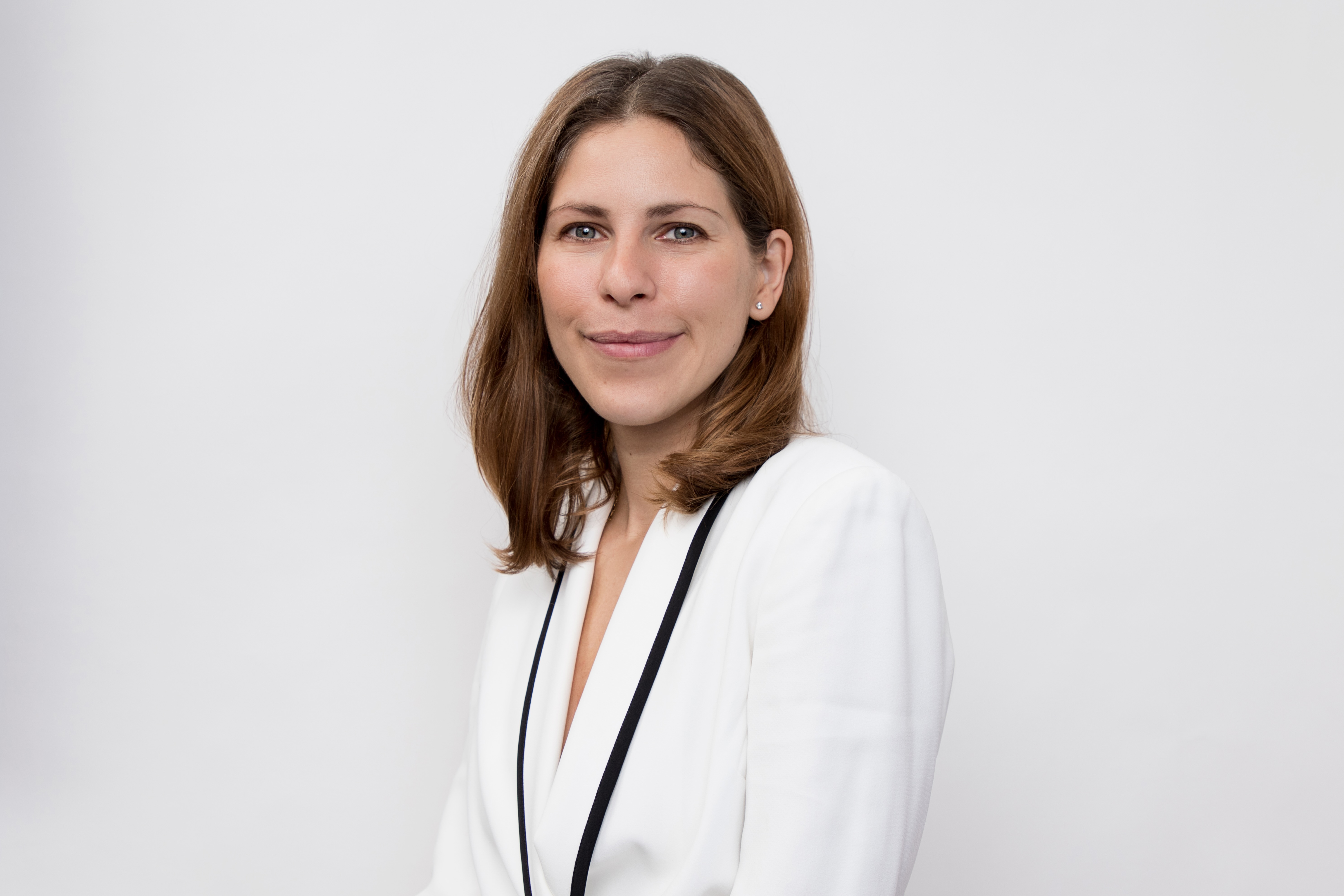 Professional portrait of a medical practitioner with brown hair wearing a white blazer with black trim against a gray background