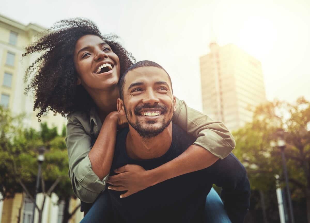 Couple sharing joyful piggyback ride in urban park with city buildings and sunset glow in background