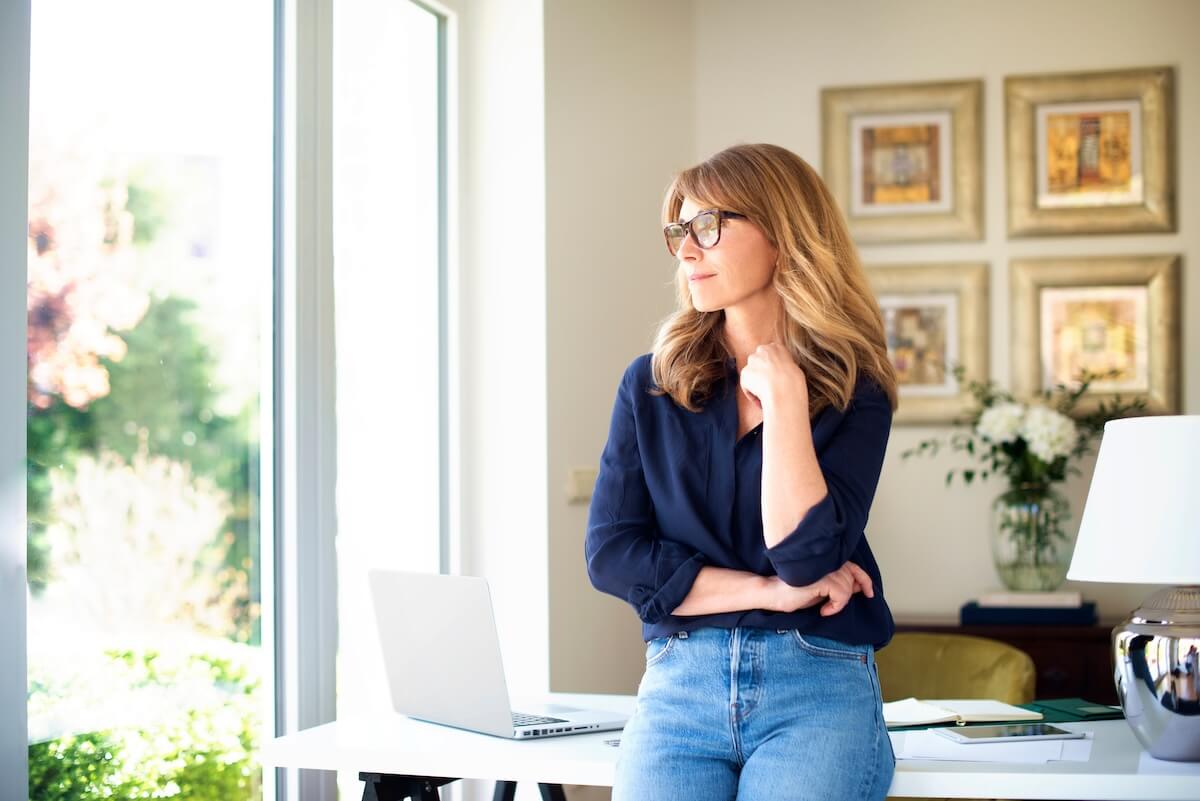 Person in glasses and navy blouse looking thoughtfully out window, home office with desk, laptop and framed art
