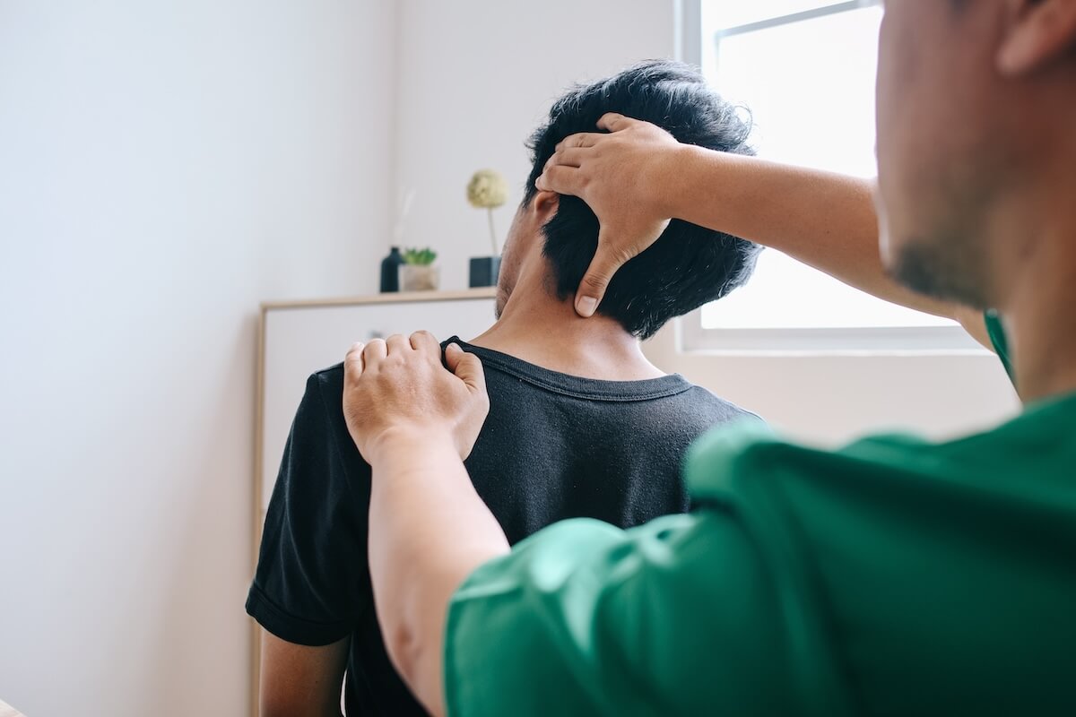 Healthcare provider in green examining patient's neck and head from behind, in bright clinical room with minimal decor